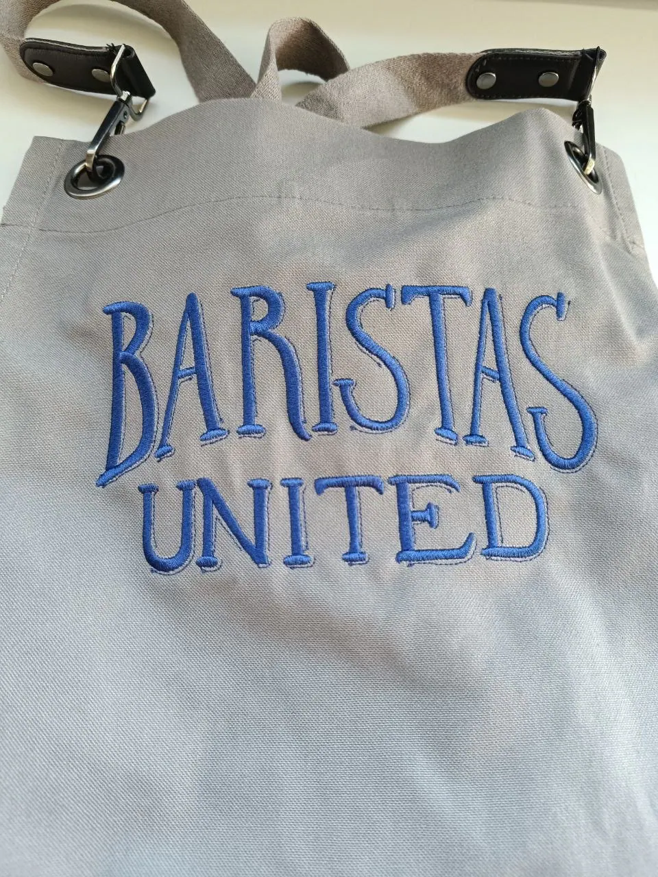 Royalblauer Baristas United Schriftzug gestickt auf hellgrauer Canvas-Schürze