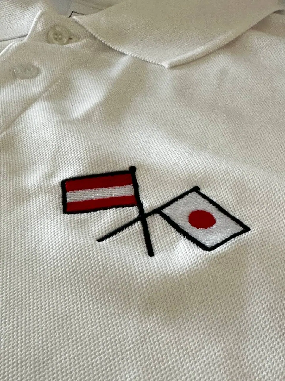 Weißes Poloshirt mit gekreuzten Flaggen Österreich und Japan gestickt – Direktstickerei rot-schwarz auf Brust