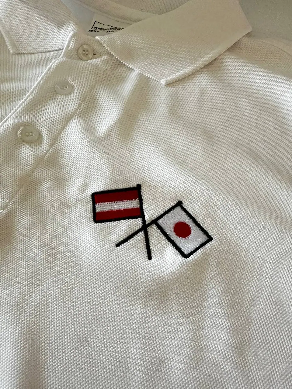 Detailaufnahme Flaggenstickerei Österreich Japan auf weißem Polo – gestickte rot-weiß und weiß-rote Flaggen