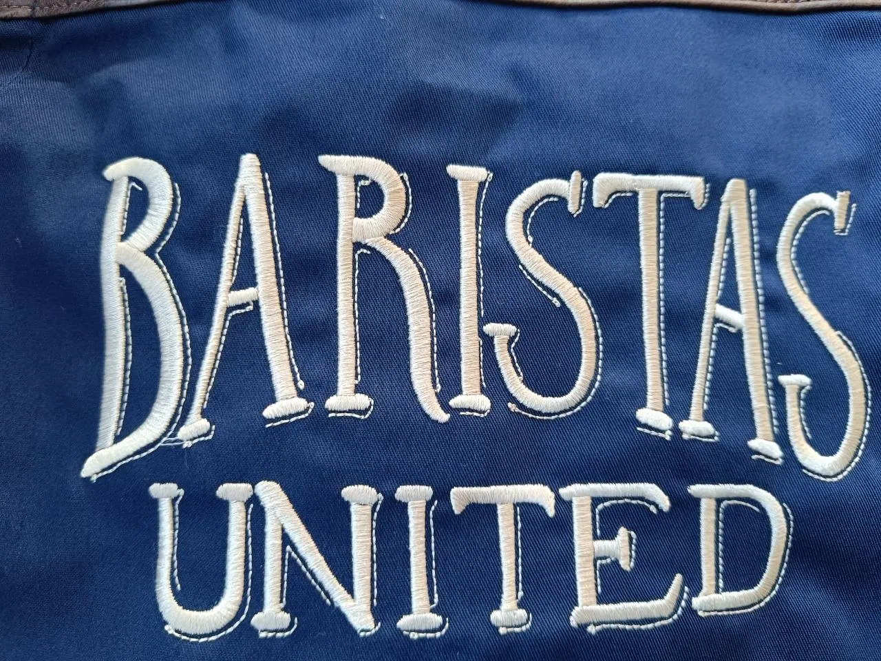 Großaufnahme Baristas United Schriftzug in beige auf navyblauer Schürze – Sticktextur